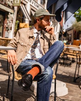 A bearded man in a tan trucker jacket and jeans seated at a café terrace, wearing black Lennertson tassel loafers with rust socks