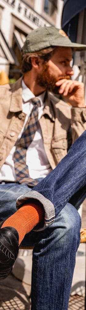 A bearded man in a tan trucker jacket and jeans seated at a café terrace, wearing black Lennertson tassel loafers with rust socks