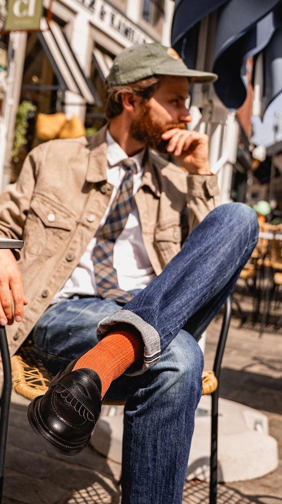 A bearded man in a tan trucker jacket and jeans seated at a café terrace, wearing black Lennertson tassel loafers with rust socks