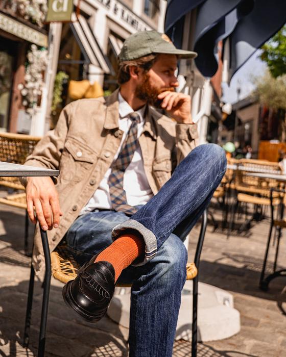 A bearded man in a tan trucker jacket and jeans seated at a café terrace, wearing black Lennertson tassel loafers with rust socks