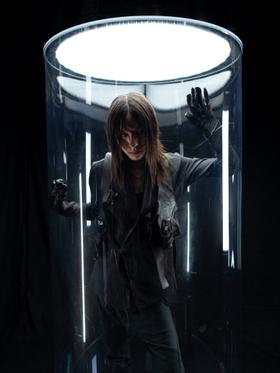 A model wearing a dark asymmetric Leon Emanuel Blanck jacket and trousers inside a lit glass cylinder
