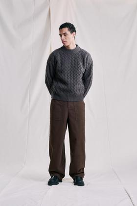 A man wearing a charcoal Libertine-Libertine cable-knit sweater with brown trousers