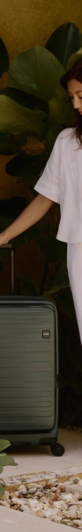A woman in a white linen shirt and trousers standing beside a dark green Lojel hard-shell suitcase