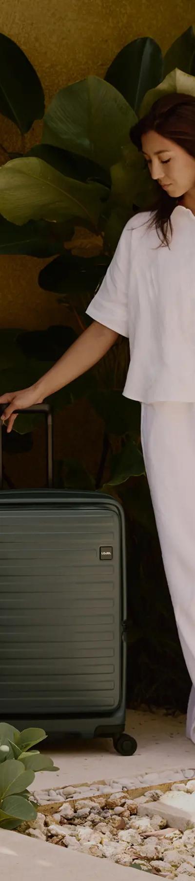 A woman in a white linen shirt and trousers standing beside a dark green Lojel hard-shell suitcase