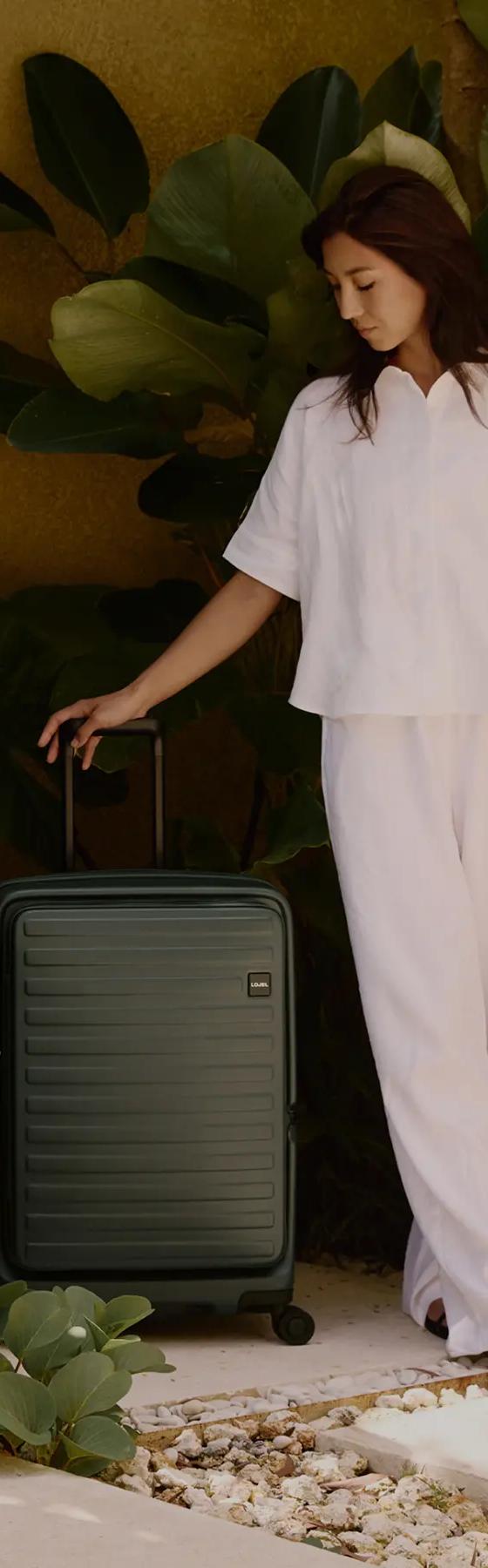 A woman in a white linen shirt and trousers standing beside a dark green Lojel hard-shell suitcase