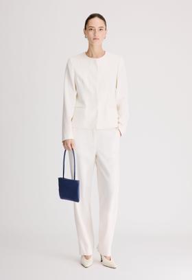 A woman wearing a cream Loulou de Saison collarless jacket and matching tailored trousers, holding a navy leather mini bag