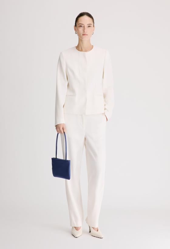 A woman wearing a cream Loulou de Saison collarless jacket and matching tailored trousers, holding a navy leather mini bag