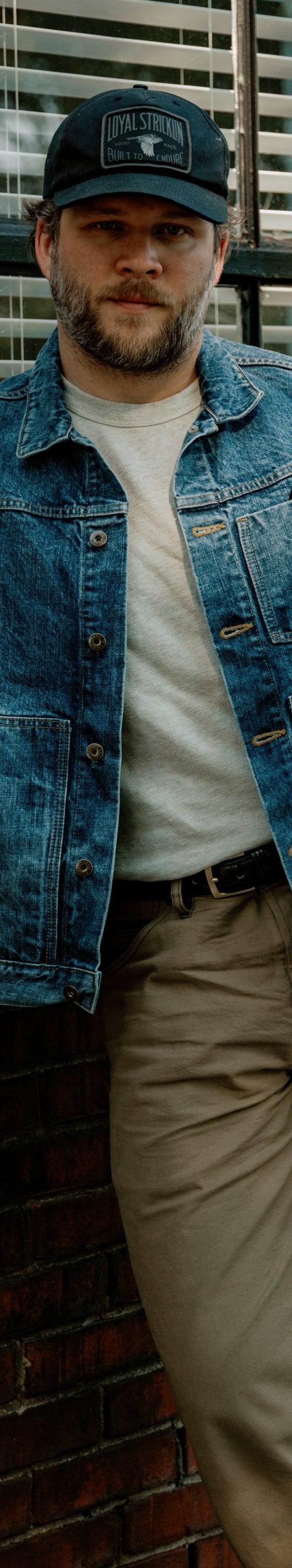 A man wearing a Loyal Stricklin denim chore jacket with a white tee and khaki trousers, leaning against a brick wall