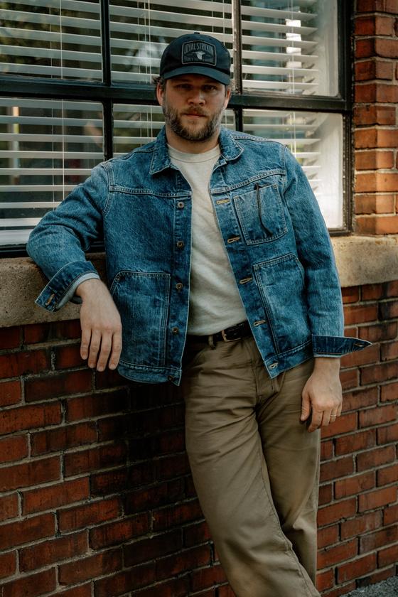 A man wearing a Loyal Stricklin denim chore jacket with a white tee and khaki trousers, leaning against a brick wall
