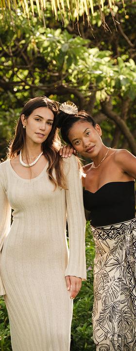 Two Mahina models in a knit dress and printed wrap skirt among palms