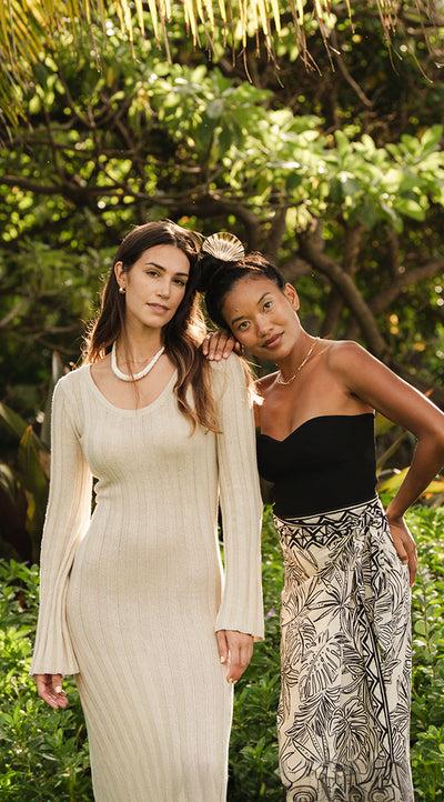 Two Mahina models in a knit dress and printed wrap skirt among palms