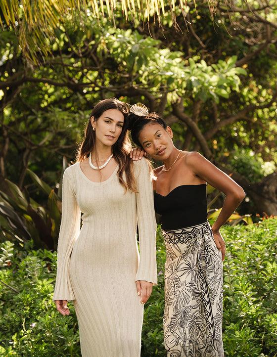 Two Mahina models in a knit dress and printed wrap skirt among palms