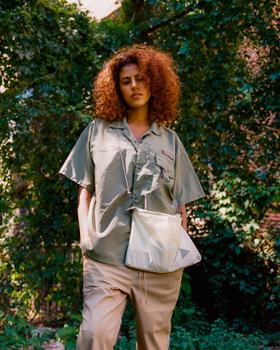 A woman wearing a sage green MANASTASH short-sleeve shirt with beige trousers and a nylon shoulder bag