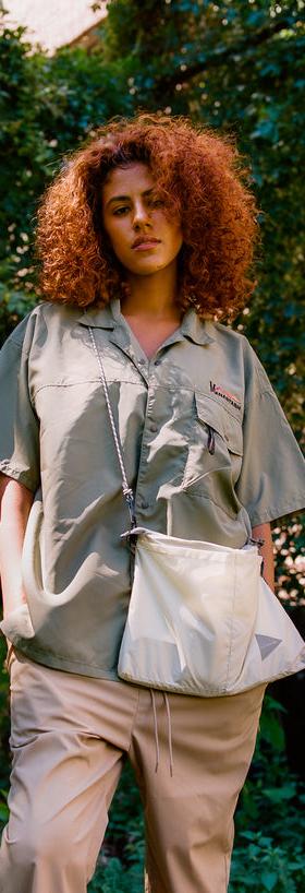 A woman wearing a sage green MANASTASH short-sleeve shirt with beige trousers and a nylon shoulder bag