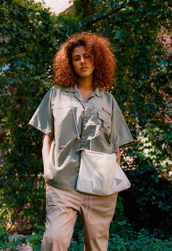A woman wearing a sage green MANASTASH short-sleeve shirt with beige trousers and a nylon shoulder bag