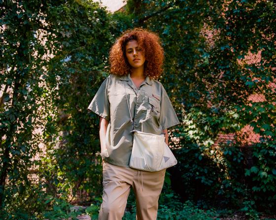 A woman wearing a sage green MANASTASH short-sleeve shirt with beige trousers and a nylon shoulder bag