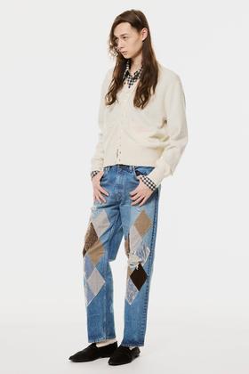 A man wearing a cream MASU cardigan over a checked shirt with argyle-patched blue jeans