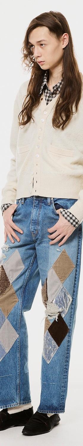 A man wearing a cream MASU cardigan over a checked shirt with argyle-patched blue jeans