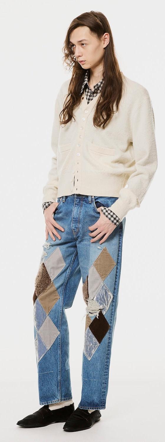 A man wearing a cream MASU cardigan over a checked shirt with argyle-patched blue jeans