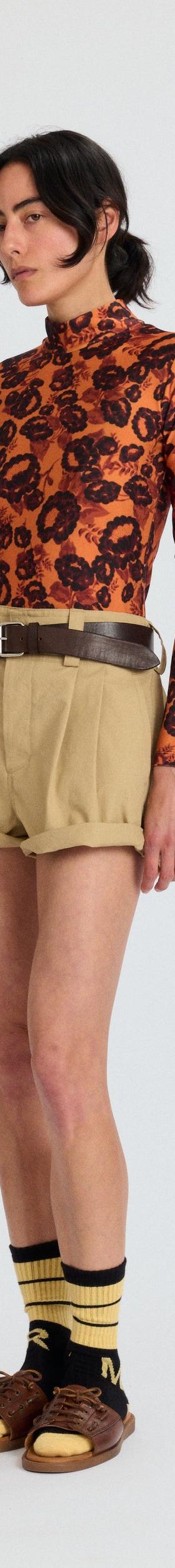 A woman wearing an orange printed Meryll Rogge turtleneck with khaki pleated shorts and brown sandals