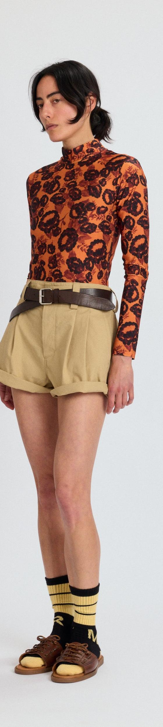 A woman wearing an orange printed Meryll Rogge turtleneck with khaki pleated shorts and brown sandals