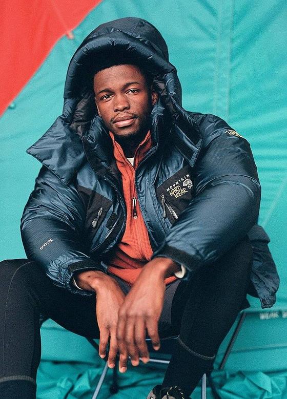 A man wearing a navy Mountain Hardwear expedition down parka over an orange fleece, seated in front of a teal tent