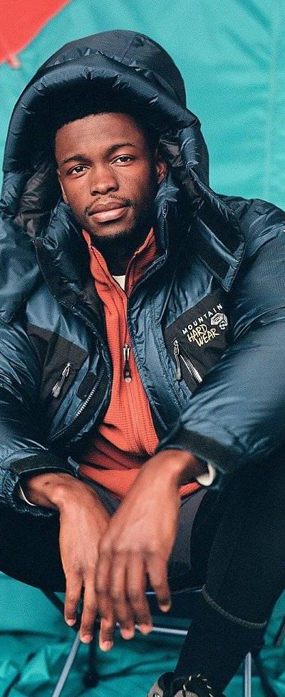 A man wearing a navy Mountain Hardwear expedition down parka over an orange fleece, seated in front of a teal tent