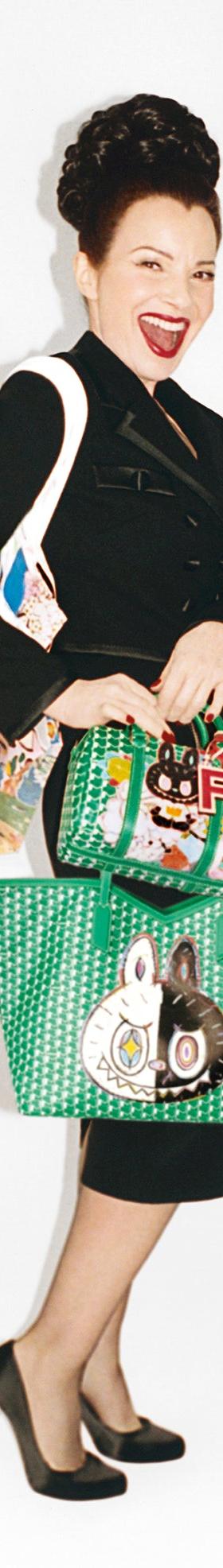 A woman in a black skirt suit and heels carrying green monogram-pattern Moynat tote and top-handle bags