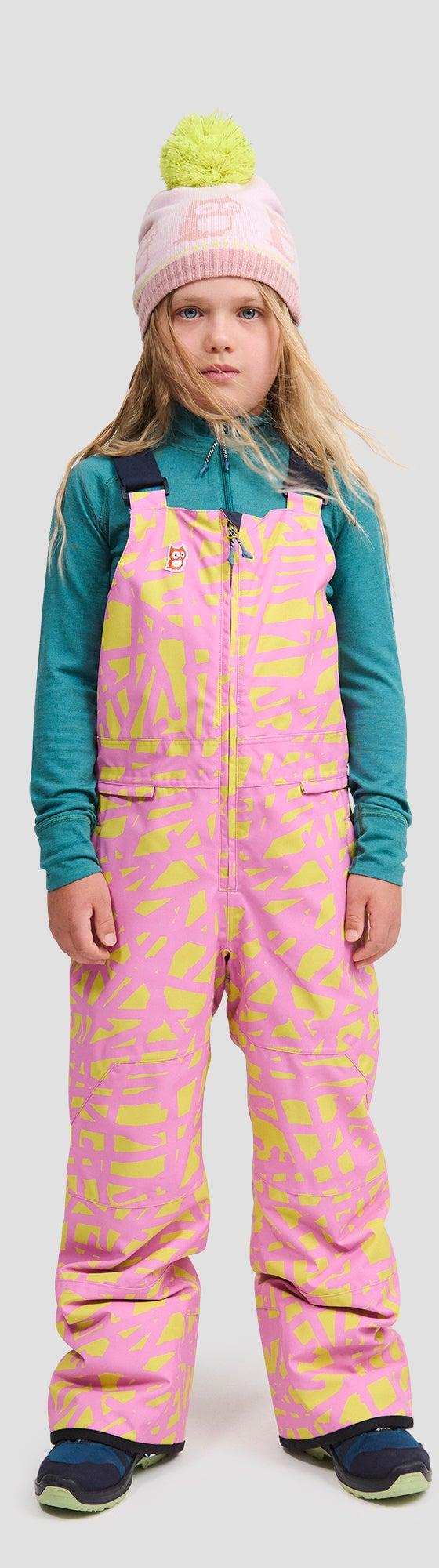 A girl wearing a pink and yellow printed namuk snow bib with a teal base layer and pink pom-pom beanie