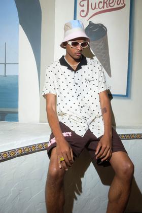 A man wearing a polka-dot camp shirt, brown shorts and a pastel bucket hat, seated outside an ice cream parlour