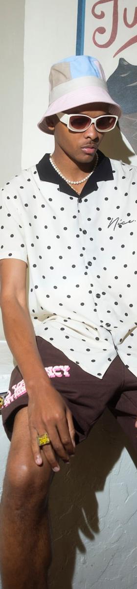 A man wearing a polka-dot camp shirt, brown shorts and a pastel bucket hat, seated outside an ice cream parlour