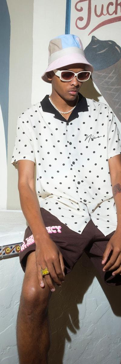 A man wearing a polka-dot camp shirt, brown shorts and a pastel bucket hat, seated outside an ice cream parlour
