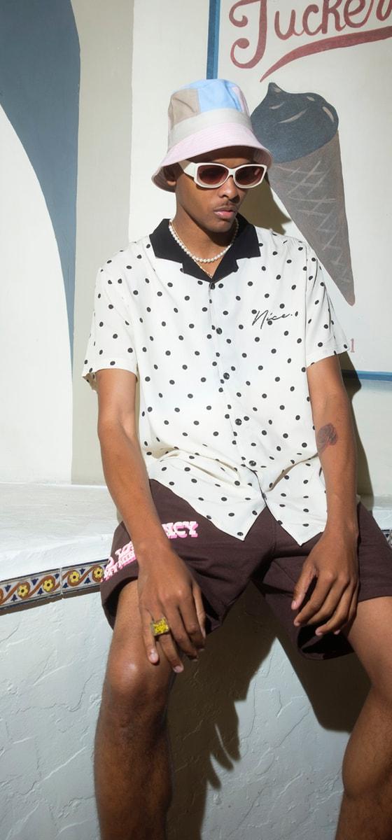 A man wearing a polka-dot camp shirt, brown shorts and a pastel bucket hat, seated outside an ice cream parlour