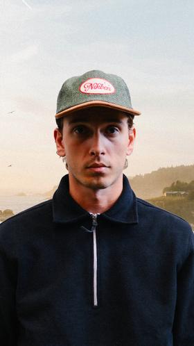 A man wearing a green herringbone Nikben cap with a suede brim and a navy half-zip sweatshirt