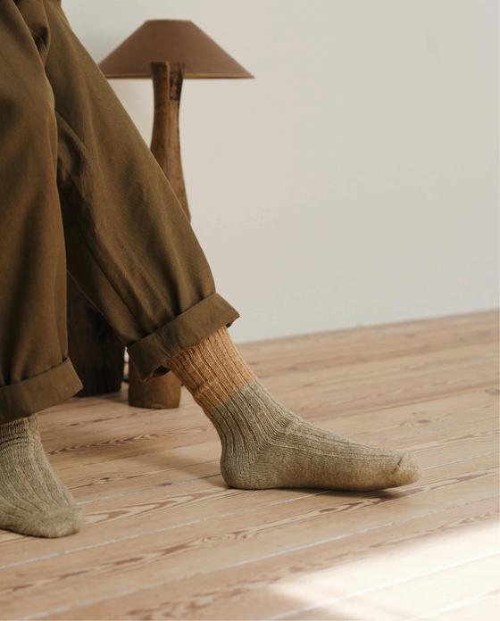 A person wearing olive ribbed NISHIGUCHI KUTSUSHITA socks with cuffed khaki trousers on a wooden floor