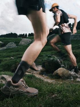 Two runners wearing NNormal trail running shoes and black shorts crossing a grassy mountain stream