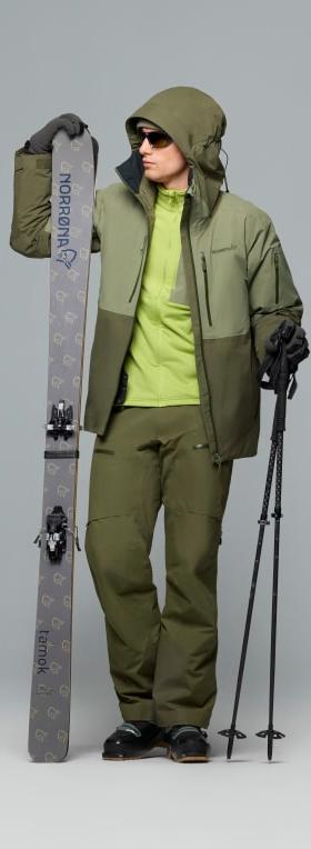 A man wearing an olive Norrøna Gore-Tex ski shell jacket and matching shell trousers, holding skis and poles