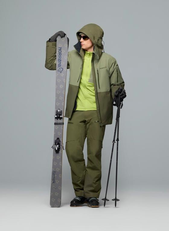 A man wearing an olive Norrøna Gore-Tex ski shell jacket and matching shell trousers, holding skis and poles
