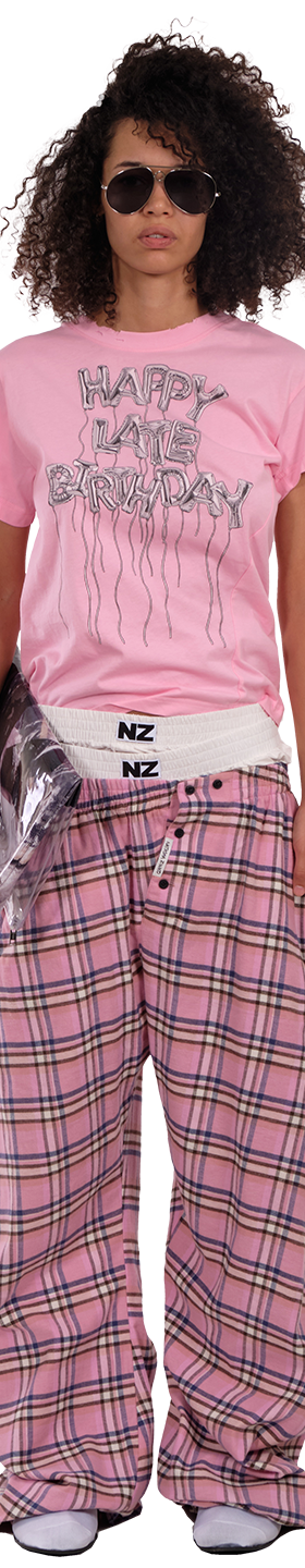 A woman wearing a pink NZKidzzz slogan T-shirt with pink checked wide-leg trousers