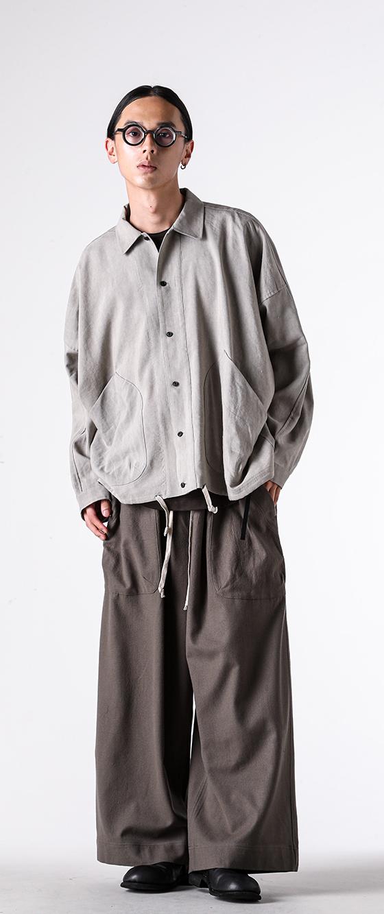 A man wearing a grey O-Project overshirt with wide brown trousers