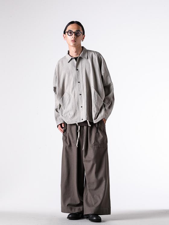 A man wearing a grey O-Project overshirt with wide brown trousers