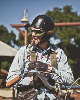 A man in a chain-stitched Old Crow Speed Shop chambray work shirt and open-face helmet with goggles, seated on a vintage motorcycle
