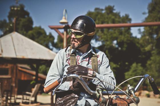 A man in a chain-stitched Old Crow Speed Shop chambray work shirt and open-face helmet with goggles, seated on a vintage motorcycle