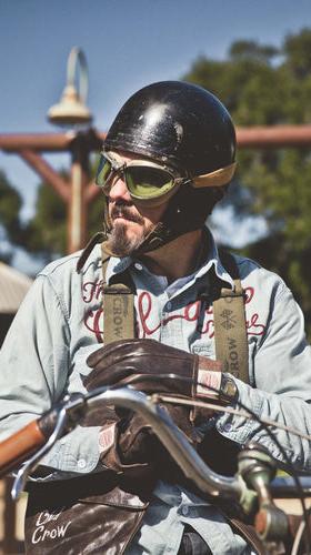 A man in a chain-stitched Old Crow Speed Shop chambray work shirt and open-face helmet with goggles, seated on a vintage motorcycle