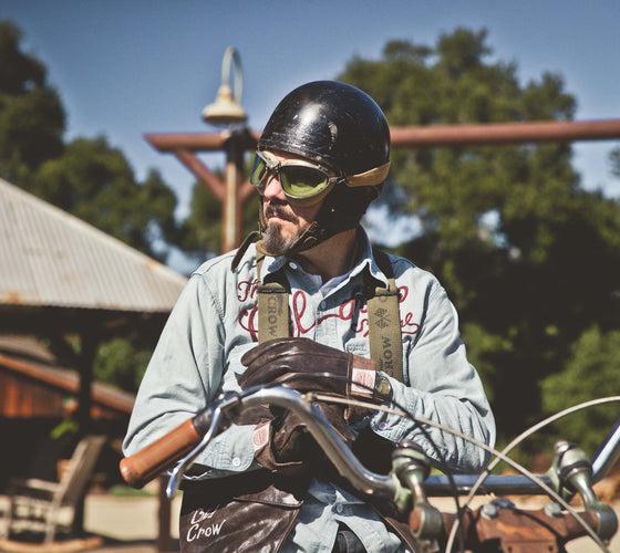A man in a chain-stitched Old Crow Speed Shop chambray work shirt and open-face helmet with goggles, seated on a vintage motorcycle