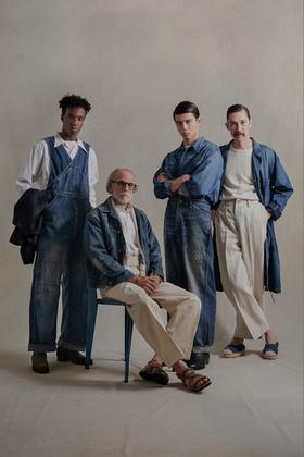 Four models in faded indigo denim overalls, chore jackets and cream trousers against a warm studio backdrop
