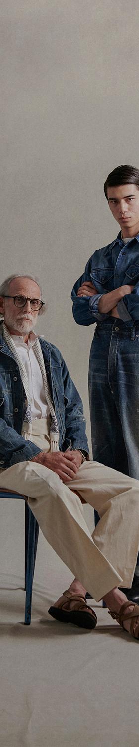 Four models in faded indigo denim overalls, chore jackets and cream trousers against a warm studio backdrop