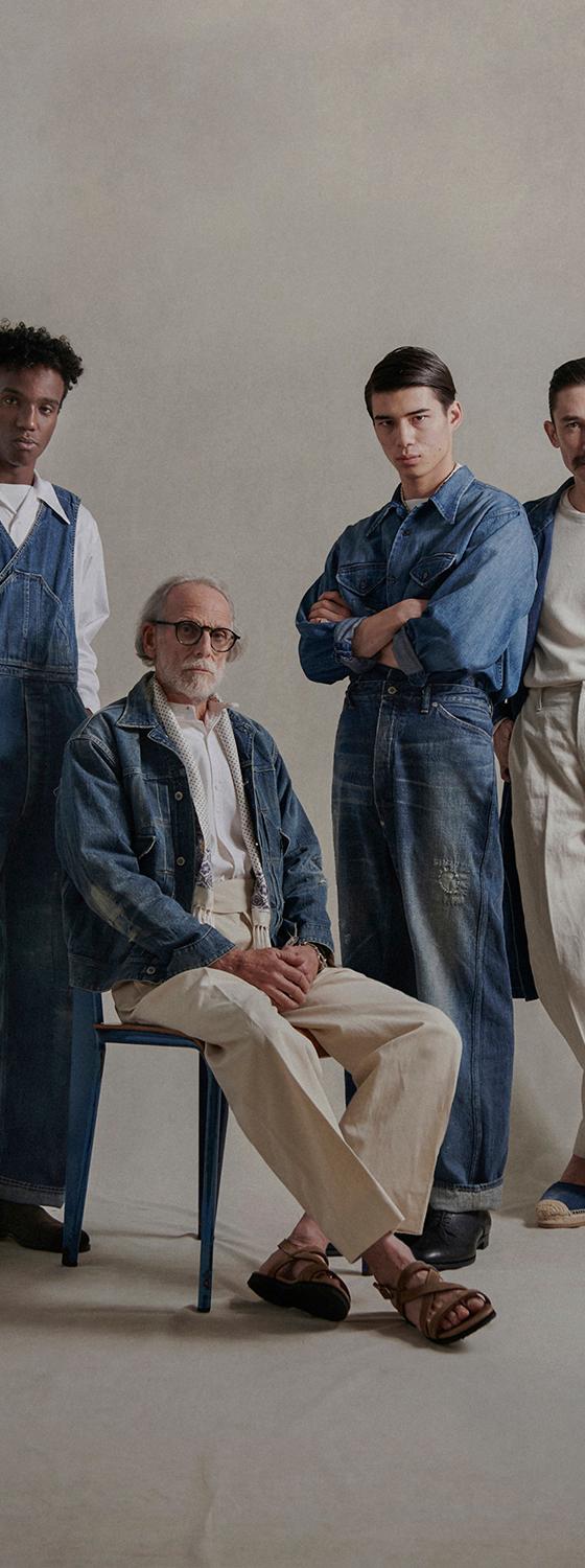 Four models in faded indigo denim overalls, chore jackets and cream trousers against a warm studio backdrop