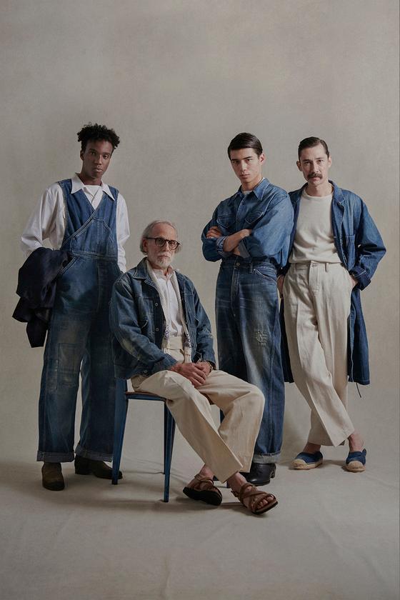 Four models in faded indigo denim overalls, chore jackets and cream trousers against a warm studio backdrop