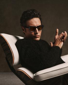 A man wearing tortoise Oliver Peoples sunglasses and a black shearling sweater, seated in a leather lounge chair